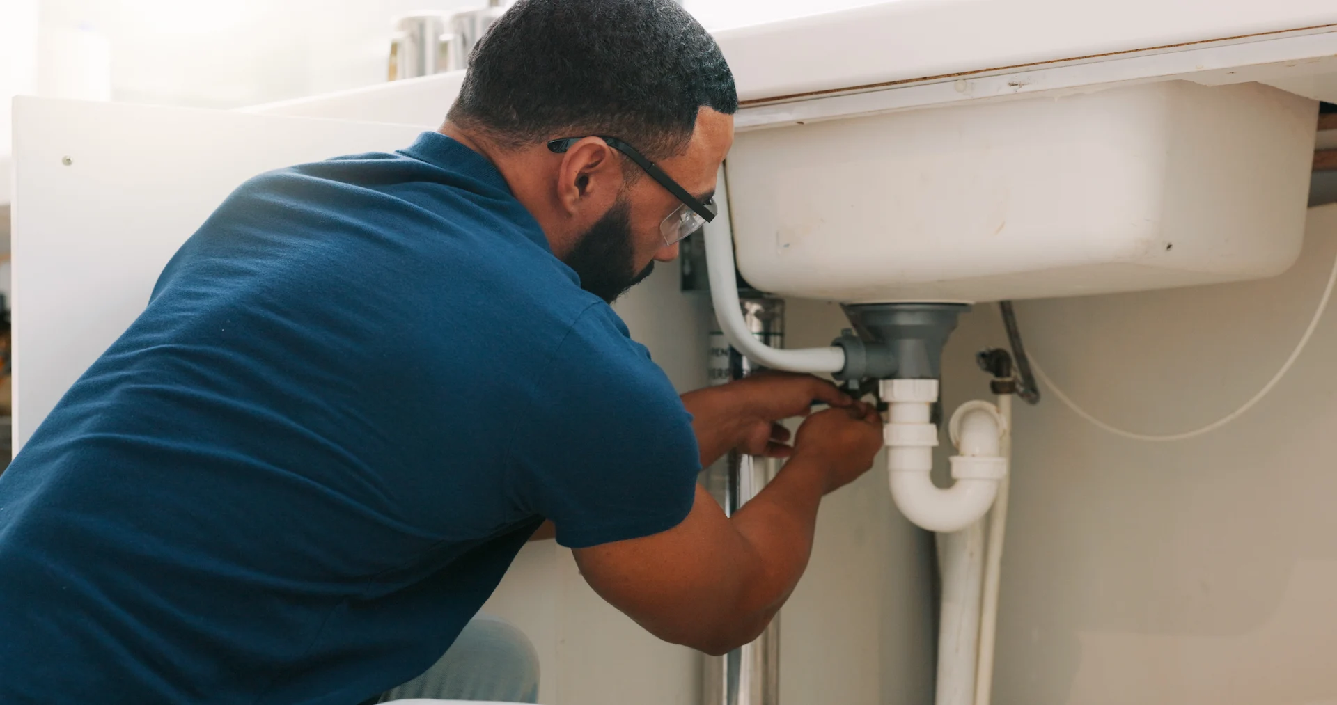 Technician in blue shirt and safety glasses installs white PVC plumbing pipes and fixtures under a bathroom sink.