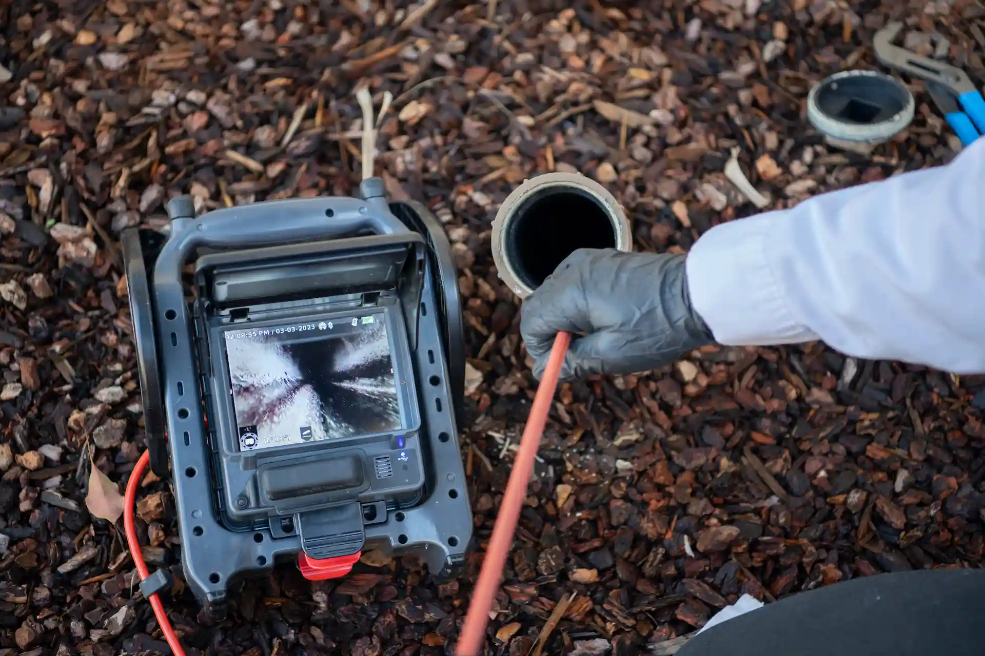 Technician holds drain inspection camera probe while monitoring sewer line video feed on handheld device screen dated 03-03-2023.