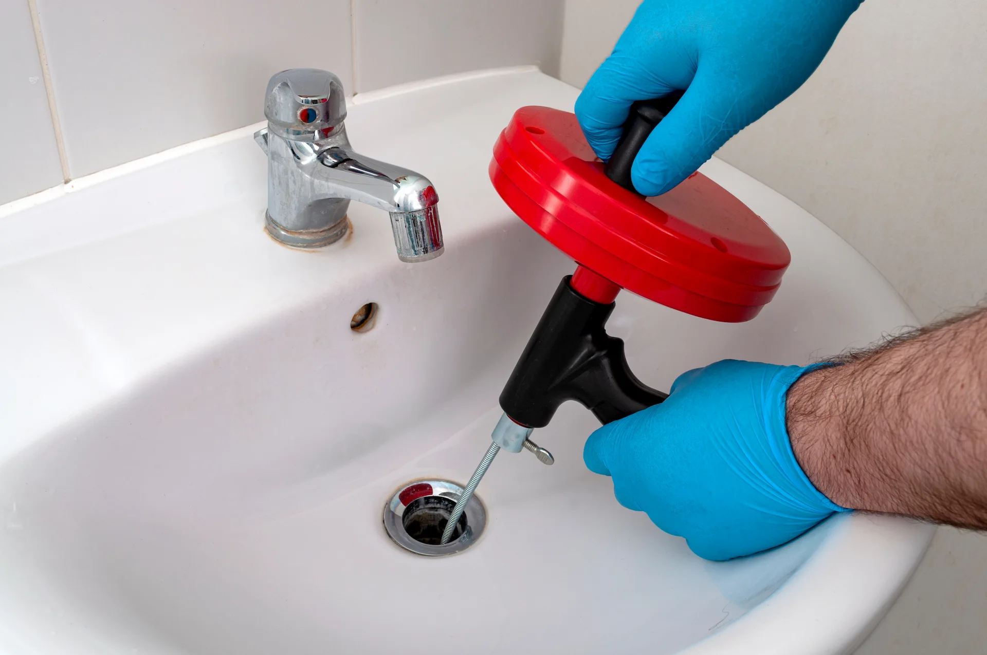 Plumber wearing blue gloves uses a red plumbing auger to clear a clogged bathroom sink drain.