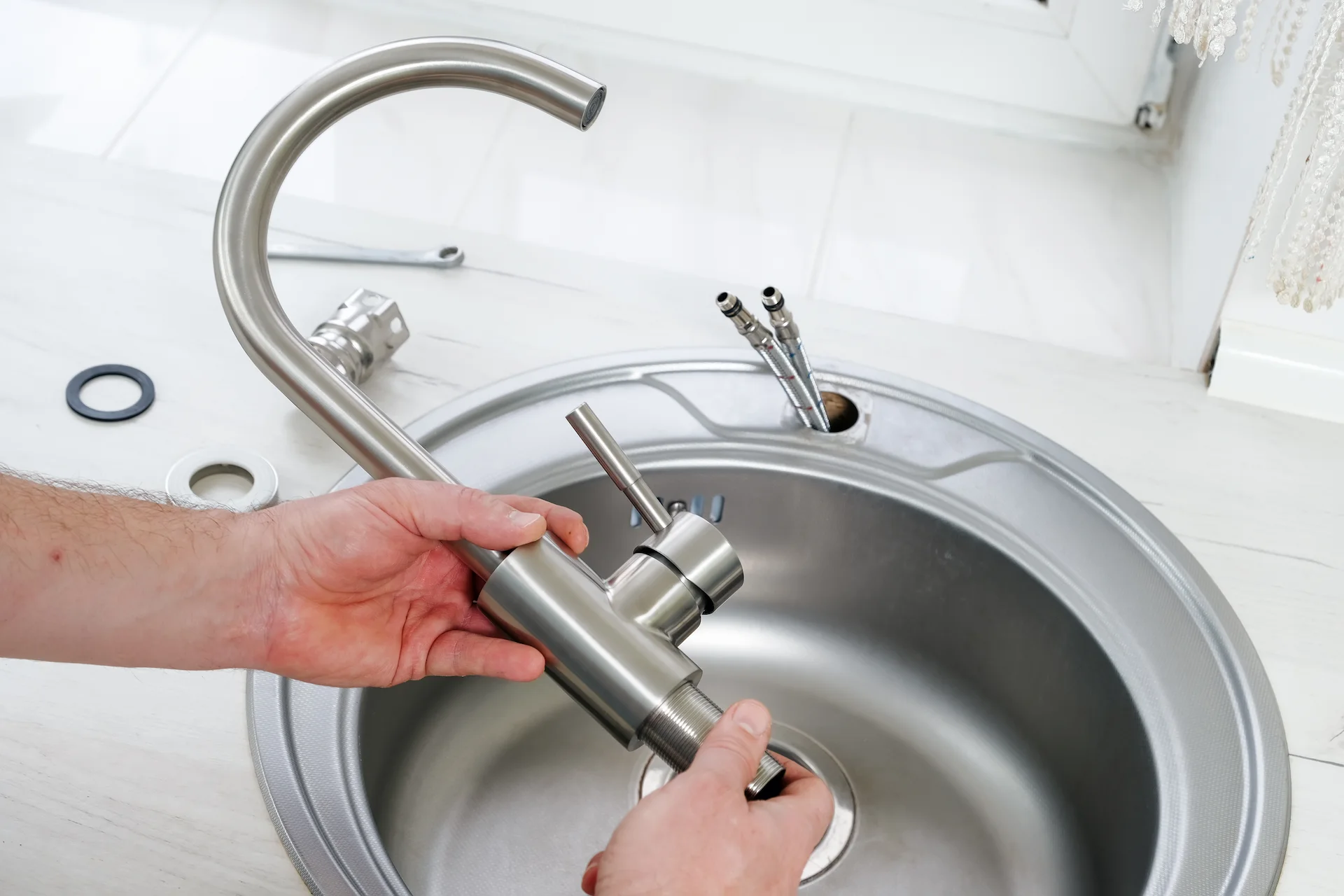 Hands installing a stainless steel kitchen faucet into a sink with plumbing components visible.