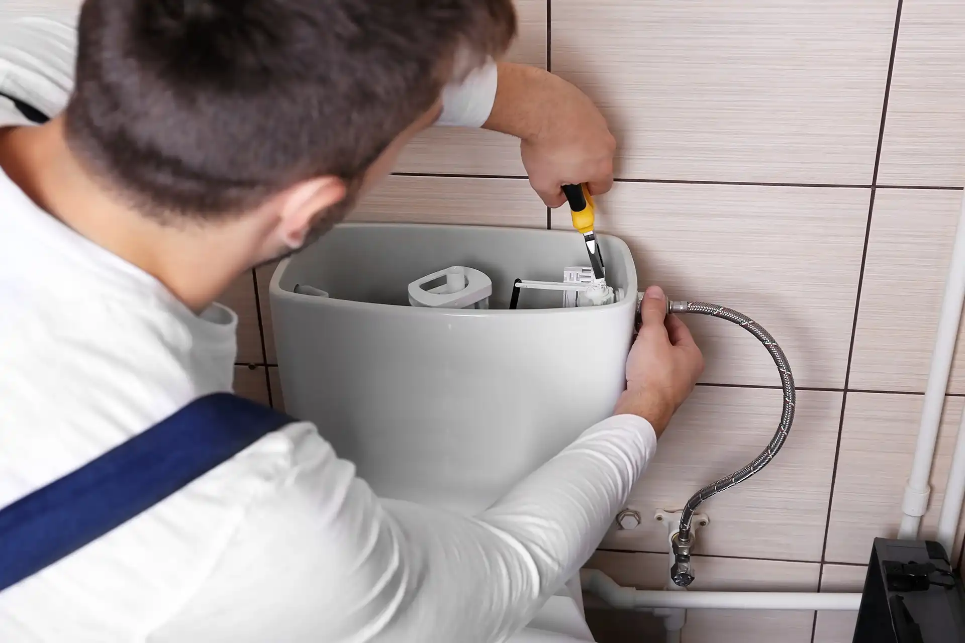 Plumber installing flexible braided metal water supply line to toilet tank fixture mounted on bathroom wall tile.